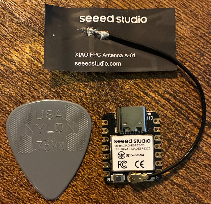 A Seeed Studio XIAO ESP32C3 next to a grey Jim Dunlop nylon 0.73 mm guitar pick, with the XIAO FPC antenna still on its backing card